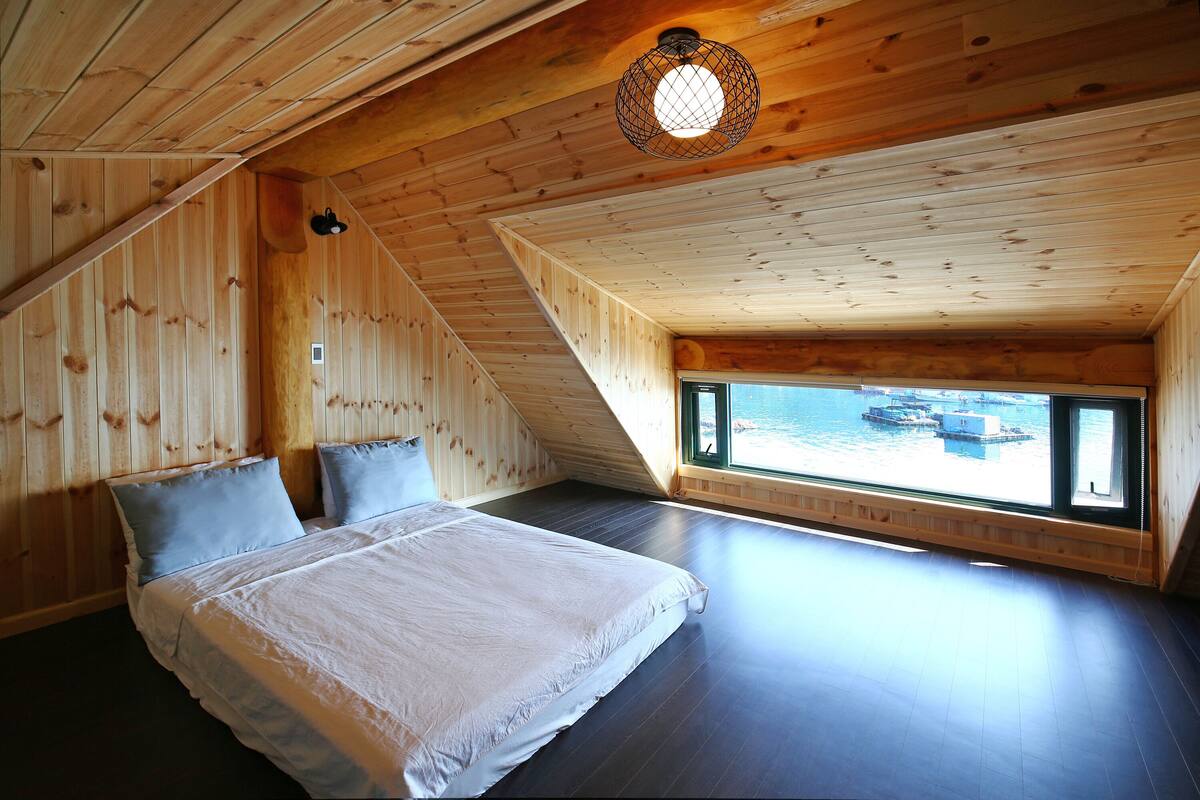 Ein Schlafzimmer mit einem großen Bett, Holzwänden, einer schrägen Decke und einem Fenster mit Blick auf das Wasser.
