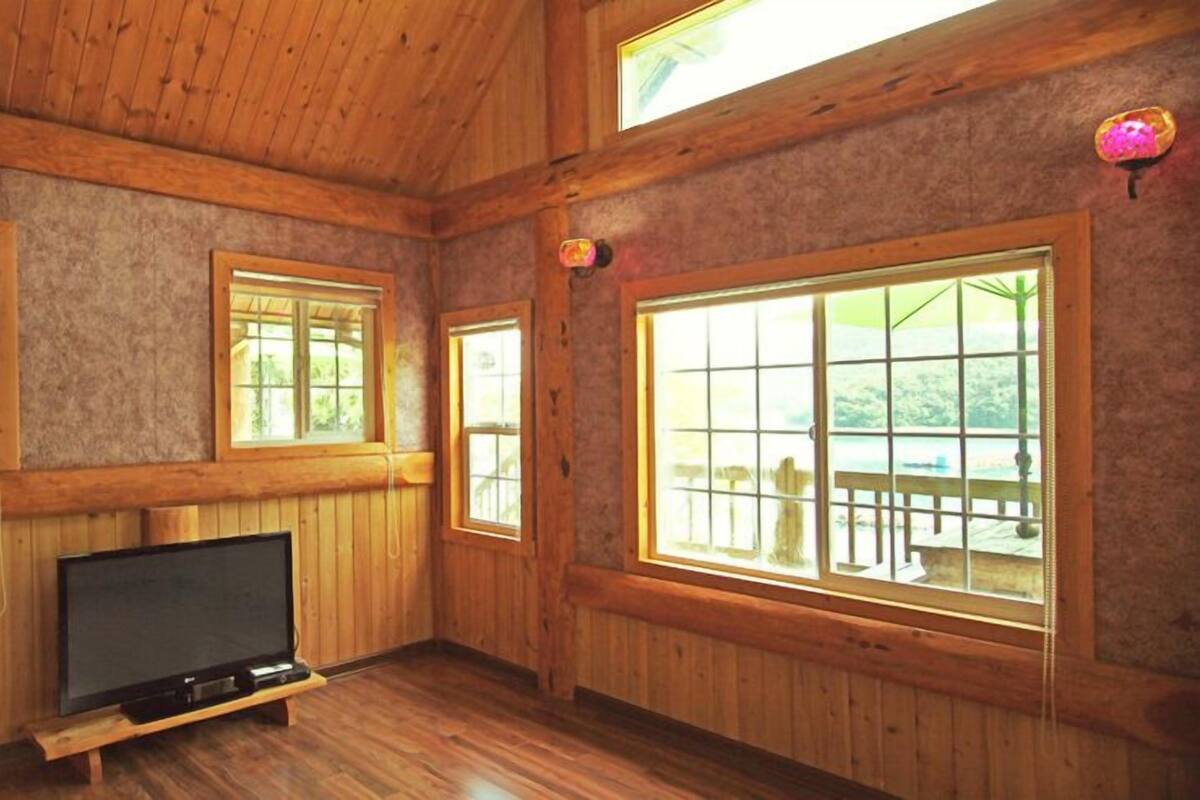 Ein Zimmer mit Holzvertäfelung, einem Fernseher auf einem Schrank, großen Fenstern und Blick auf eine Terrasse.