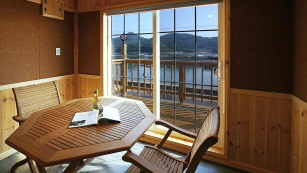 A wooden interior with a table and chairs, a window with a view of a body of water and mountains, and a ceiling fan. - Tongyeong Daore Pension (Tongyeong)