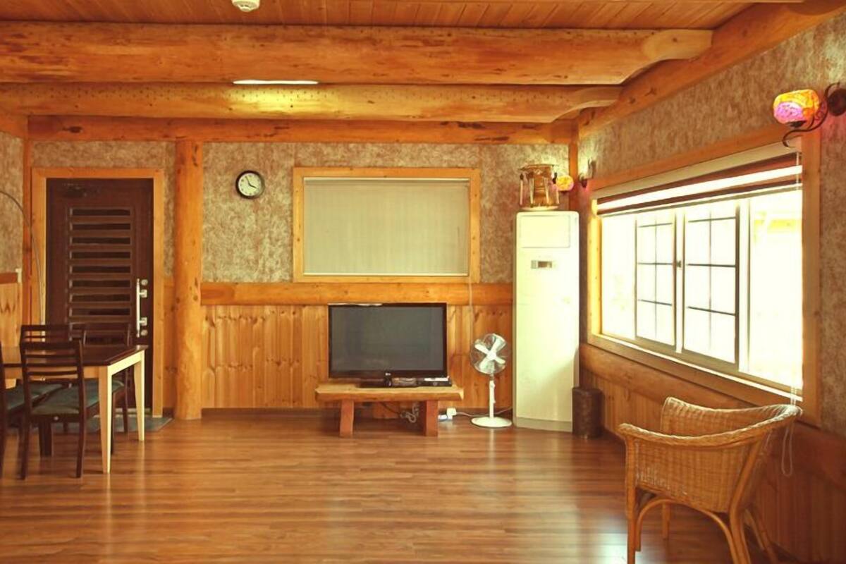 Ein gemütliches Zimmer mit Holzwänden und Holzdielen, einem Fernseher auf einem Schrank, einem Esstisch mit Stühlen, einem Ventilator und einem Fenster mit Jalousien.