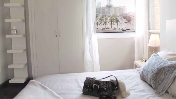 A hotel room with a bed, a window with a cityscape view, a white wardrobe, and a bedside table with a lamp. - Incredible apartment-central-sea view-near the beach-Free Wifi. (Barcelona)