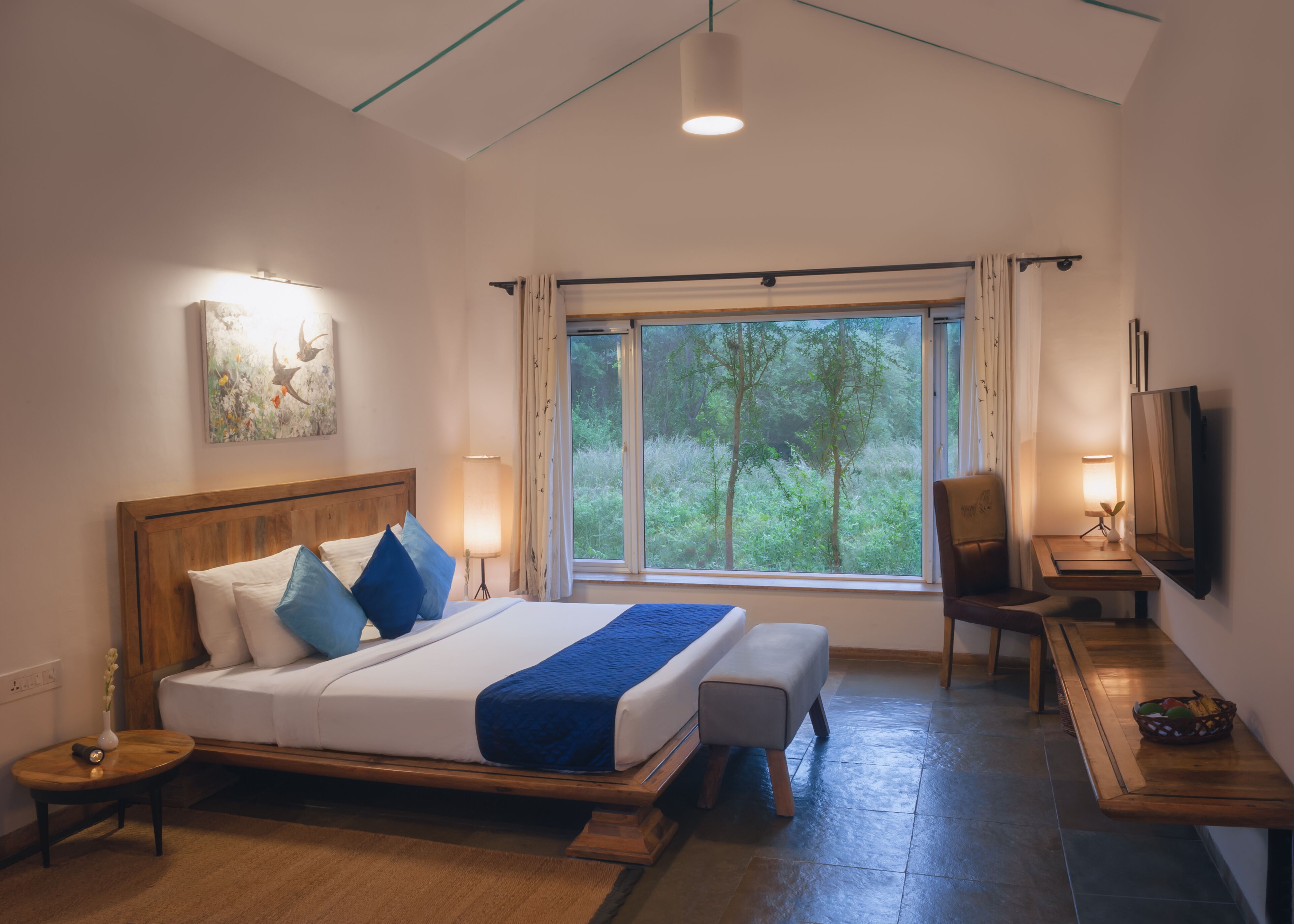 A bedroom with a large bed, bedside lamps, a desk with a chair, a television, and a window with a view of greenery.
