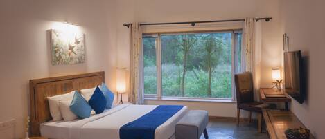 A bedroom with a large bed, bedside lamps, a desk with a chair, a television, and a window with a view of greenery.