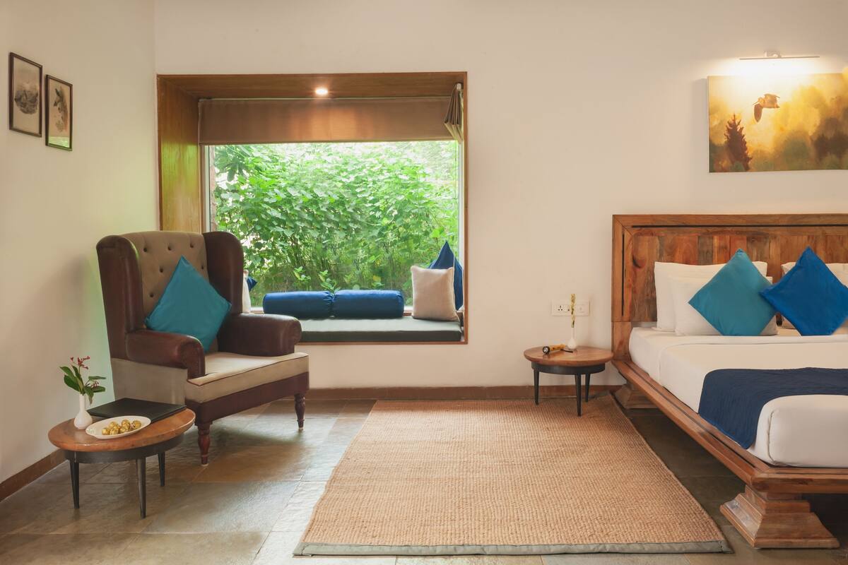 A room with a large window, a bed with blue pillows, a chair with a cushion, a small table with a plant, and a painting on the wall.