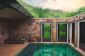 A stone-walled room with a pool, a lounge area, and large windows offering a view of greenery.