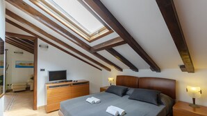 Un dormitorio con tragaluz, vigas de madera, una cama con almohadas, un televisor, un aparador y una mesita de noche con lámpara.