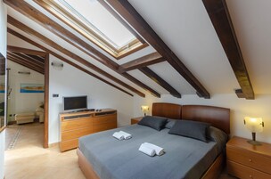Un dormitorio con tragaluz, vigas de madera, una cama con almohadas, un televisor, un aparador y una mesita de noche con lámpara.