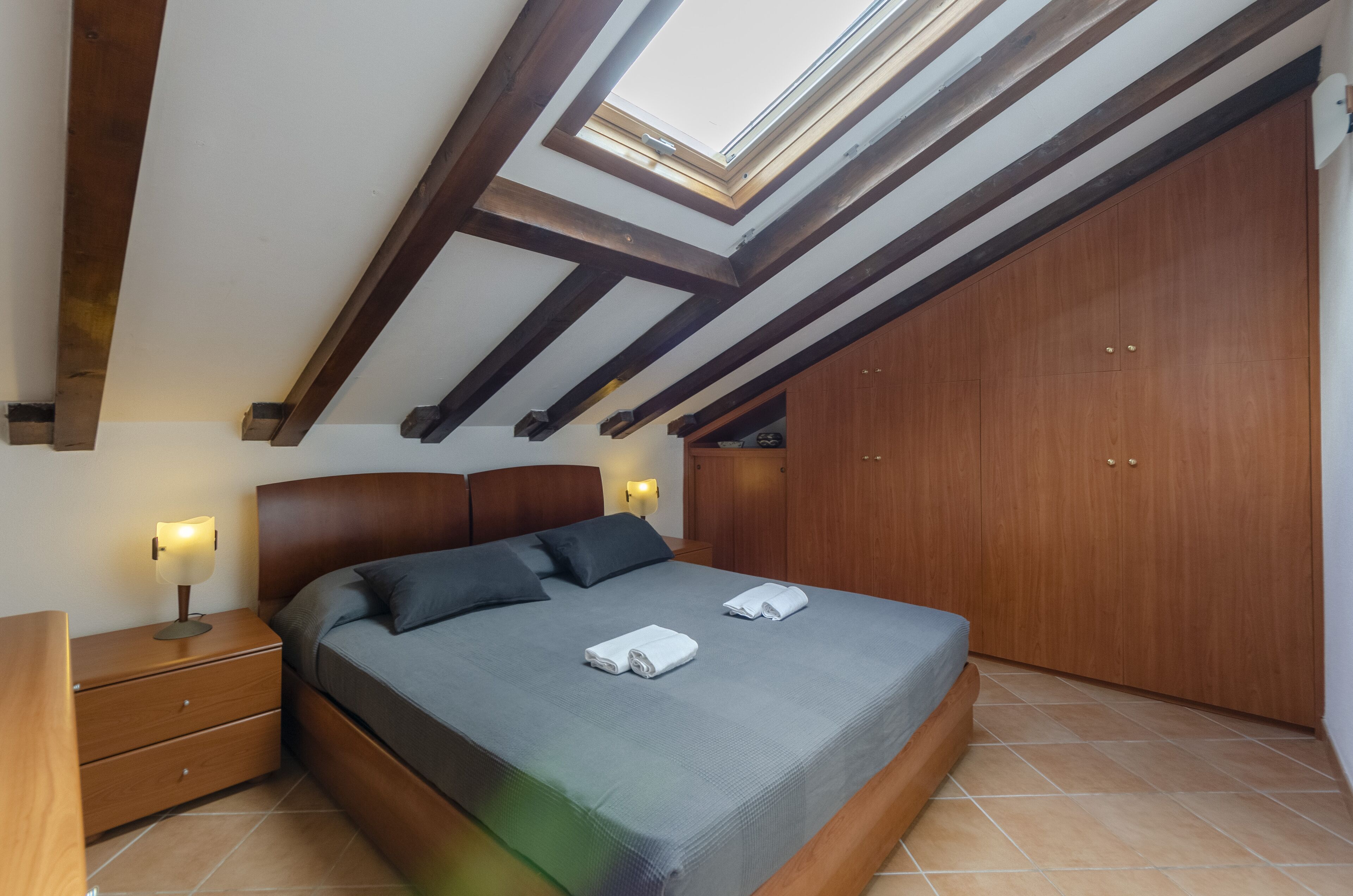 Ein Schlafzimmer mit Oberlicht, Holzbalken, einem Bett mit Kissen, einem Nachttisch und einem Kleiderschrank.