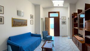 Un salon avec un canapé bleu, un fauteuil à motifs et une table basse en bois. Les murs sont ornés de tableaux encadrés, et une porte donne sur l’extérieur.