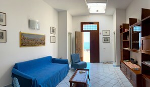 Una sala de estar con un sofá azul, un sillón estampado y una mesa de centro de madera. Hay cuadros enmarcados en las paredes y una vista al exterior a través de una puerta.