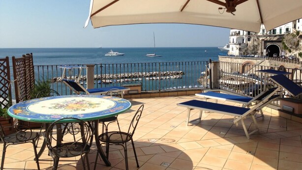 Une terrasse avec une table et des chaises, un parasol et une vue sur la mer, où l’on peut apercevoir des bateaux.