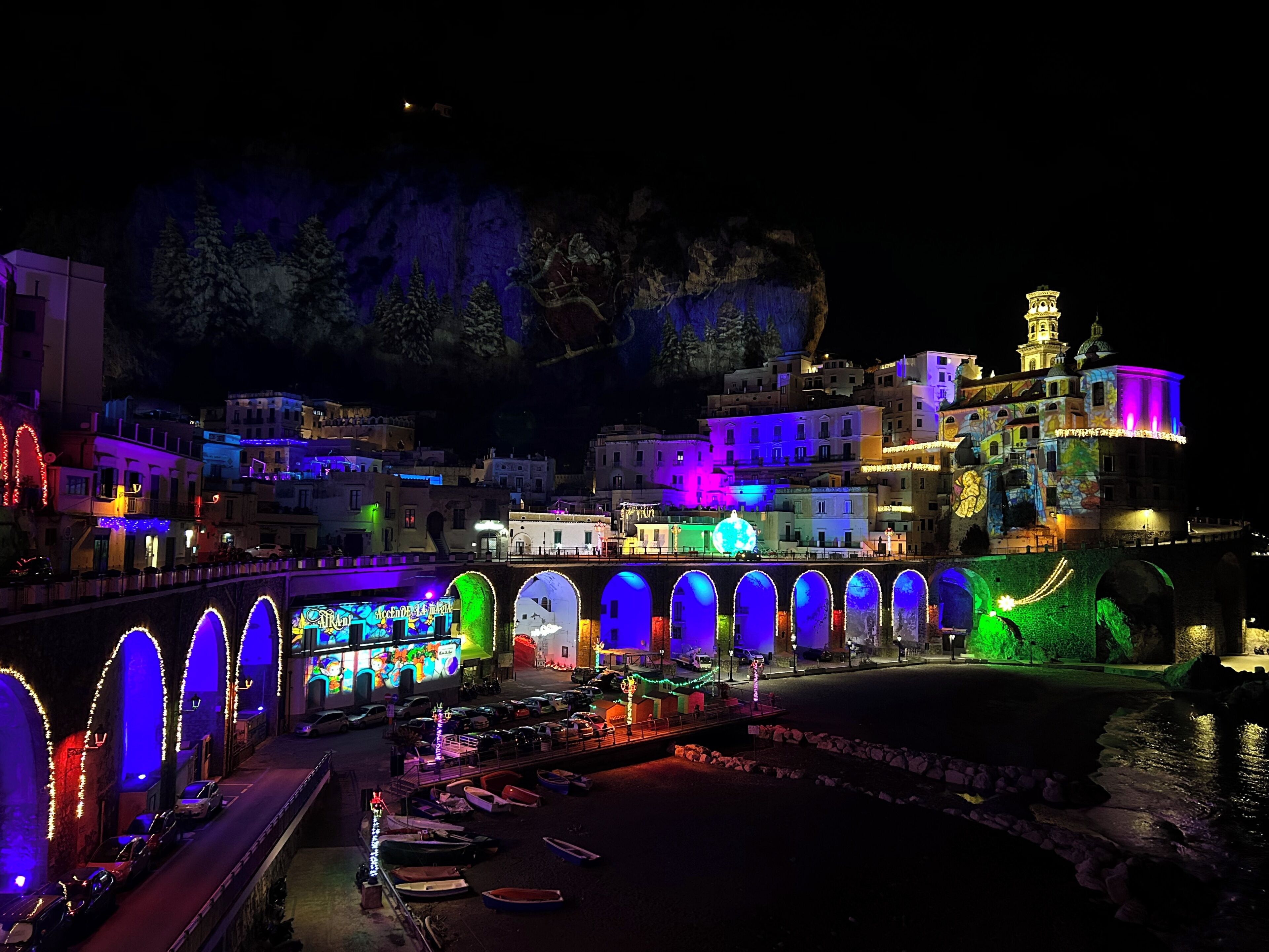 A nighttime view of a coastal town with illuminated buildings, a bridge with arches, and boats docked along the waterfront.