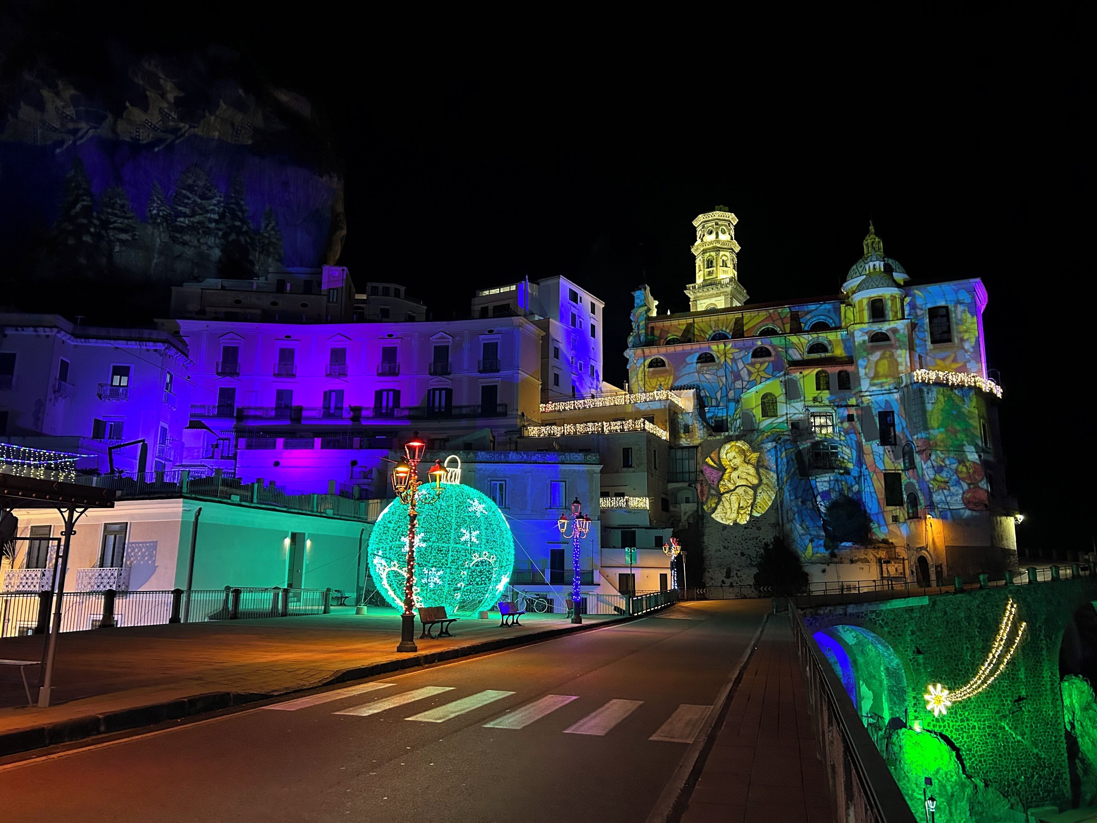 A nighttime street view with illuminated buildings, a large spherical light installation, and a bridge with decorative lights.