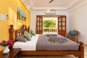A bedroom with a bed, a wooden headboard, a window with an open door, a wicker chair, and a wall-mounted air conditioner. - 10BHK Private Family Friendly Self Catered Villa - 2 min walk to Calangute beach (Calangute)