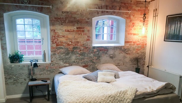 A bedroom with a brick wall, two windows, a bed with a white blanket, and a bedside table with a lamp. - Hos Mette (Borkop)