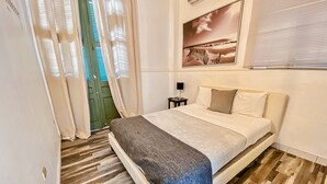 A bedroom with a bed, a nightstand, a lamp, a window with blinds, and a door with curtains. - Gladioli 1206 - That's all Puerto Rico (Ponce Pueblo)