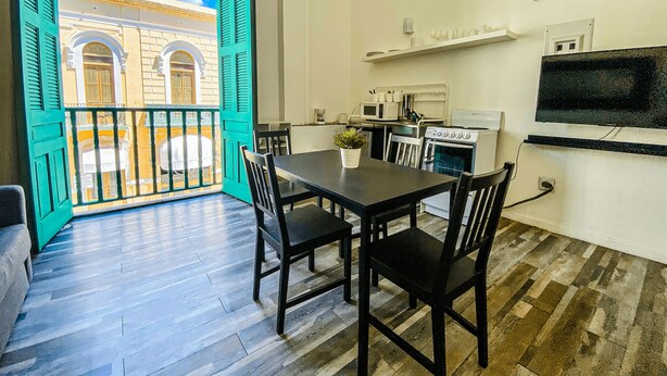 A dining area with a table and chairs, a kitchen with appliances, and a balcony with a view of a building. - Gladioli 1206 - That's all Puerto Rico (Ponce Pueblo)