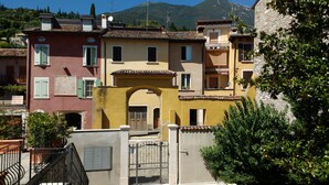 A row of Mediterranean-style buildings with arched doorways and balconies, surrounded by greenery and mountains in the background.