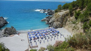 Una playa con numerosas sombrillas azules y tumbonas, costa rocosa y montañas al fondo.