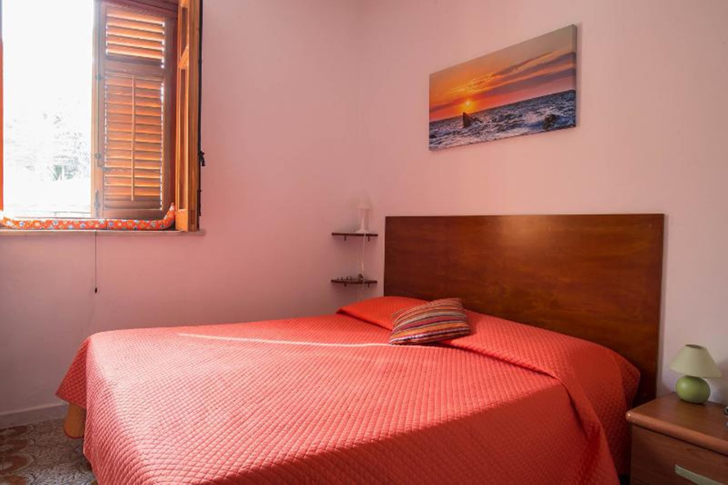 Ein Schlafzimmer mit einem Einzelbett, einem Nachttisch, einer Lampe, einem Sonnenuntergangsbild und einem Fenster mit Jalousien.