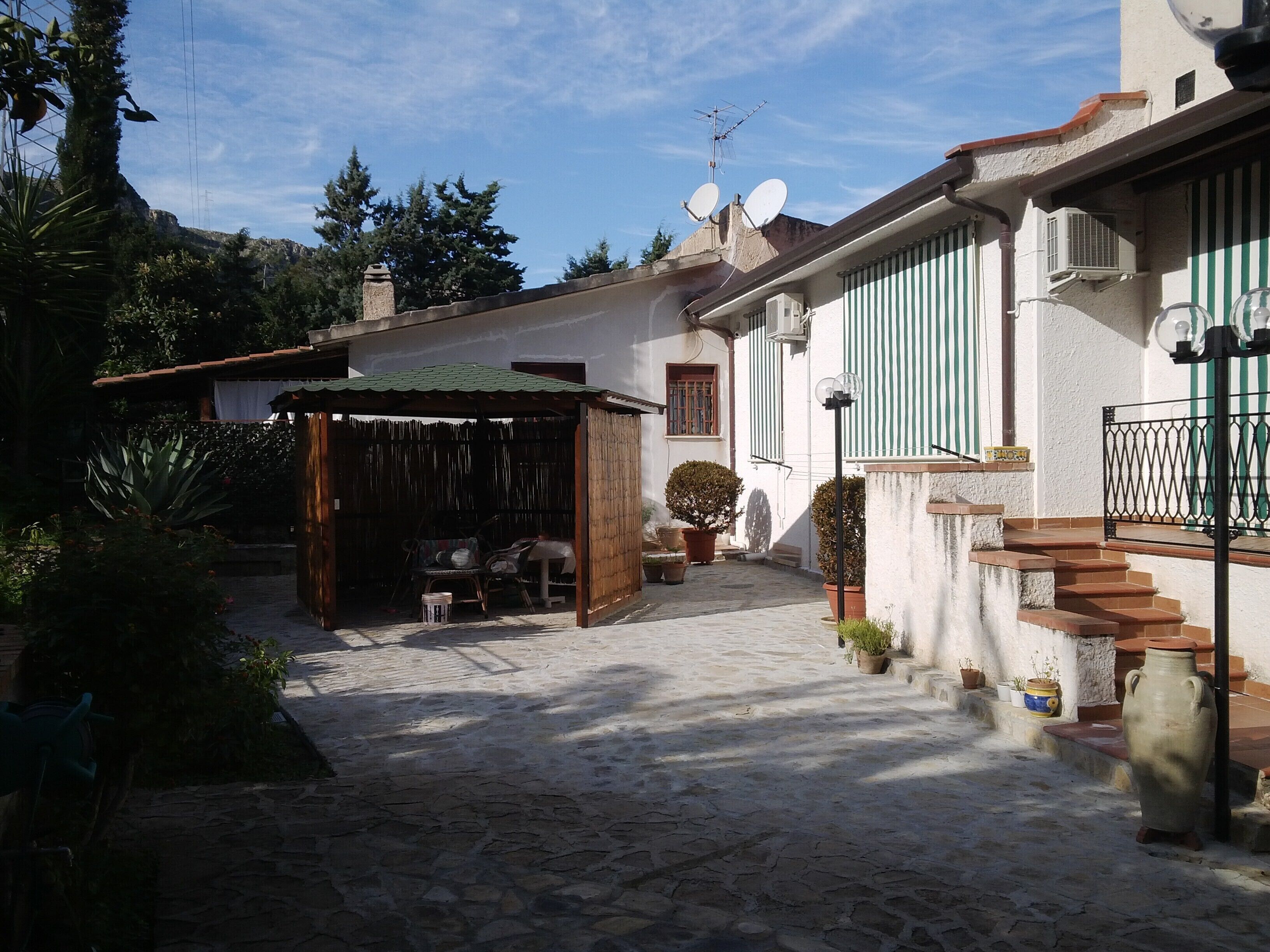 A courtyard with a covered seating area, stone flooring, potted plants, and a staircase leading to a building with striped shutters.