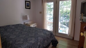 A bedroom with a large bed, a bedside table with a lamp, a window with curtains, and a view of the outside. - Just a fishing cabin in the woods... (Portland)