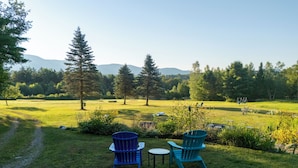 A grassy outdoor area with two blue Adirondack chairs and a small table, surrounded by trees and a distant mountain range. - Immaculate mountain cabin with awe inspiring sunrise and mountain views (Ripton)