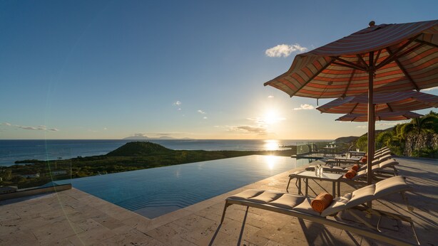 Une piscine à débordement offrant une vue sur l’océan et le coucher de soleil, des chaises longues et des parasols à rayures.