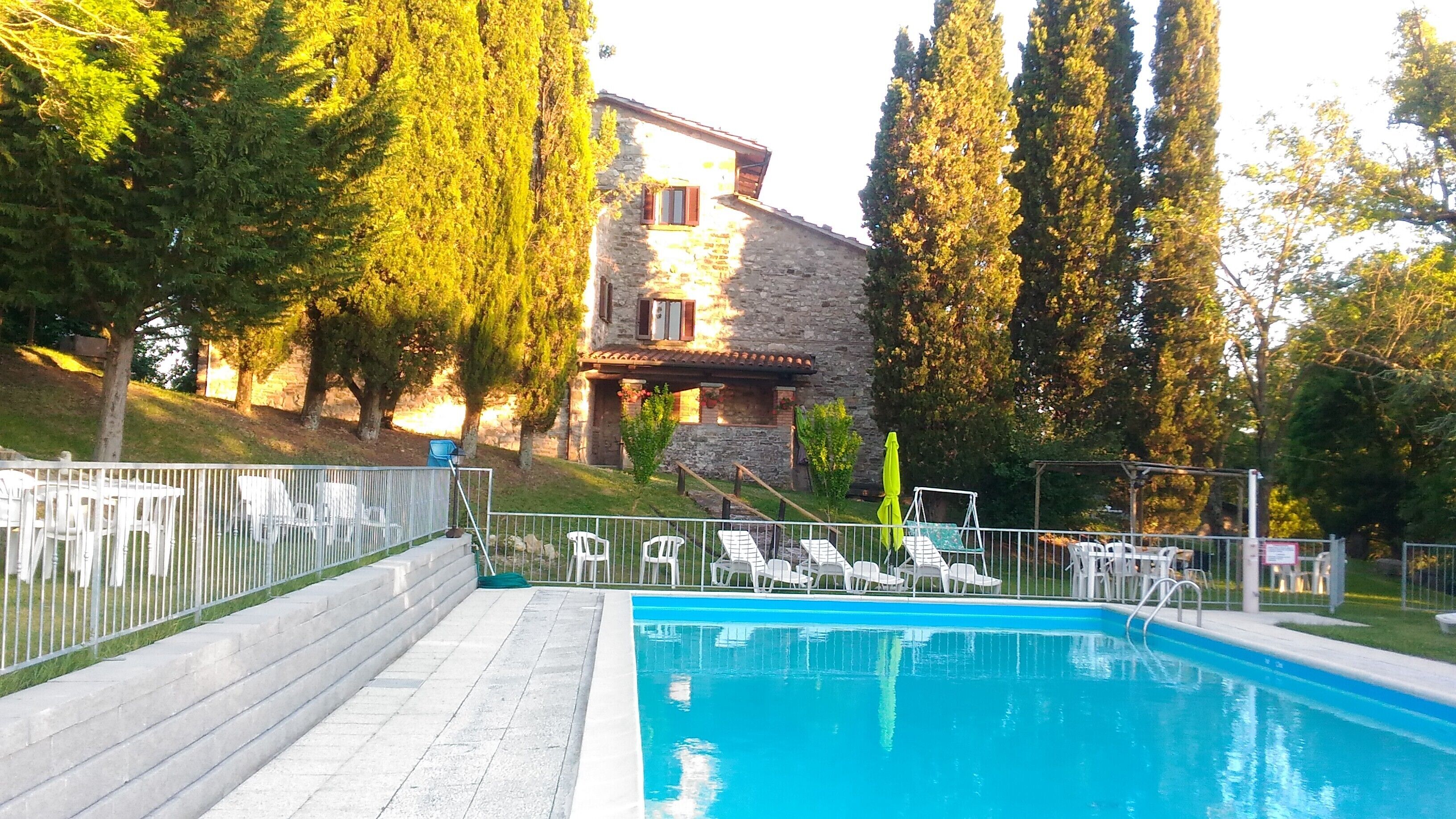 A swimming pool with lounge chairs, a stone building with a sloped roof, and tall trees in the background.