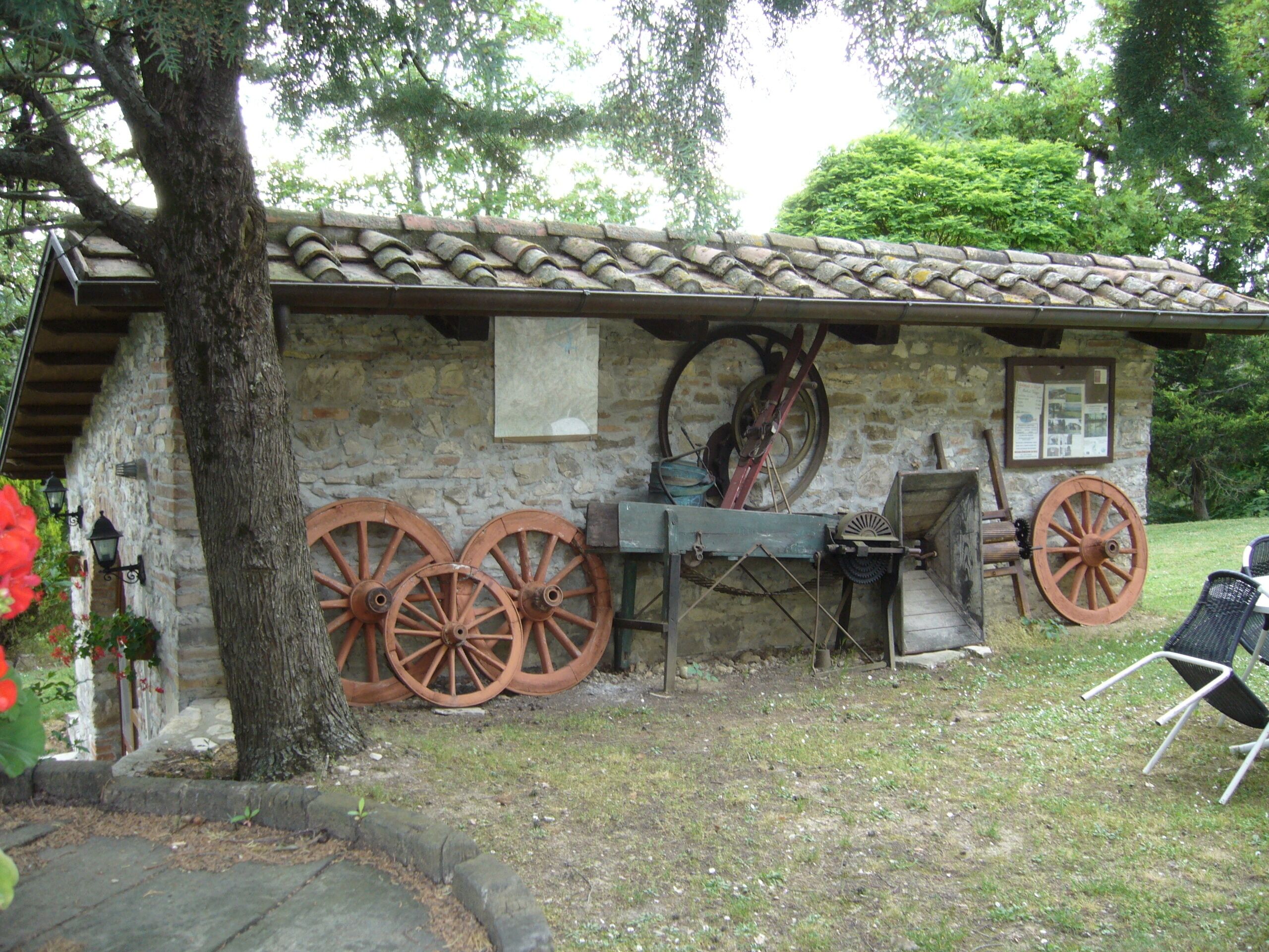 A rustic stone building with a tiled roof, large wooden wheels, and a wooden bench.