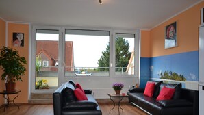 Een woonkamer met twee zwarte leren banken, een bijzettafeltje met een bloempot en grote ramen met uitzicht op een huis en bomen.