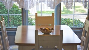 Una mesa de madera con un cuenco decorativo en el centro, frente a una ventana con cortinas de encaje y vistas al exterior con follaje.