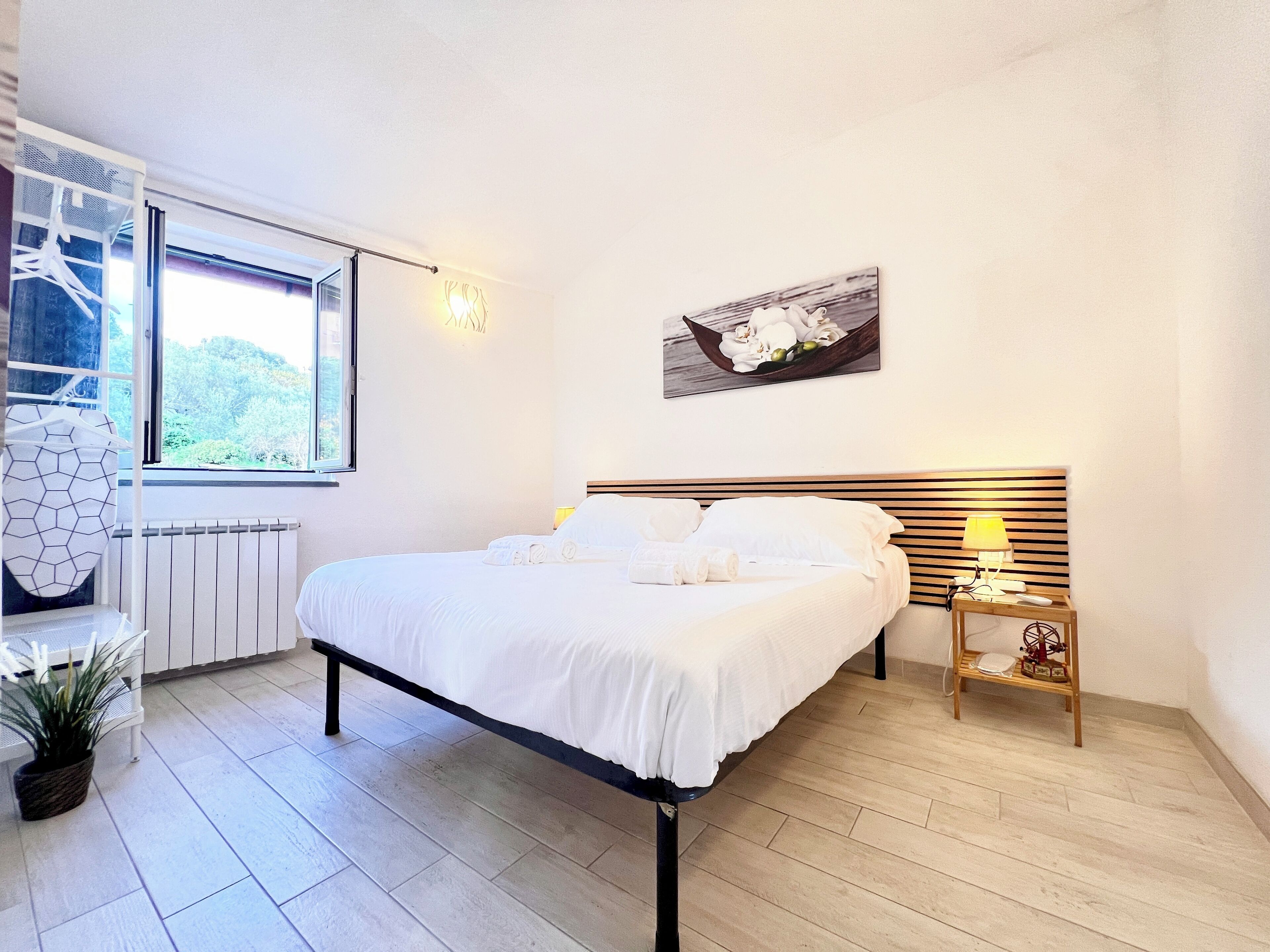 A bedroom with a bed, a nightstand, a lamp, a window, and a wall-mounted picture.