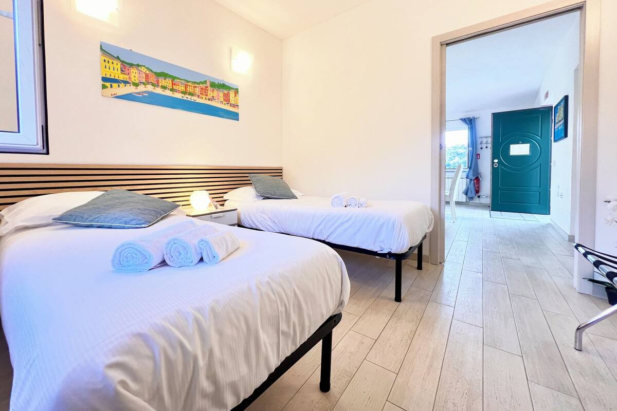 Habitación de hotel con dos camas, un cuadro de un pueblo costero, una puerta al balcón y vistas al exterior.