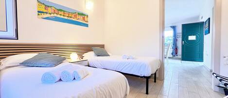 A hotel room with two beds, a painting of a coastal town, a door to the balcony, and a view of the outside.