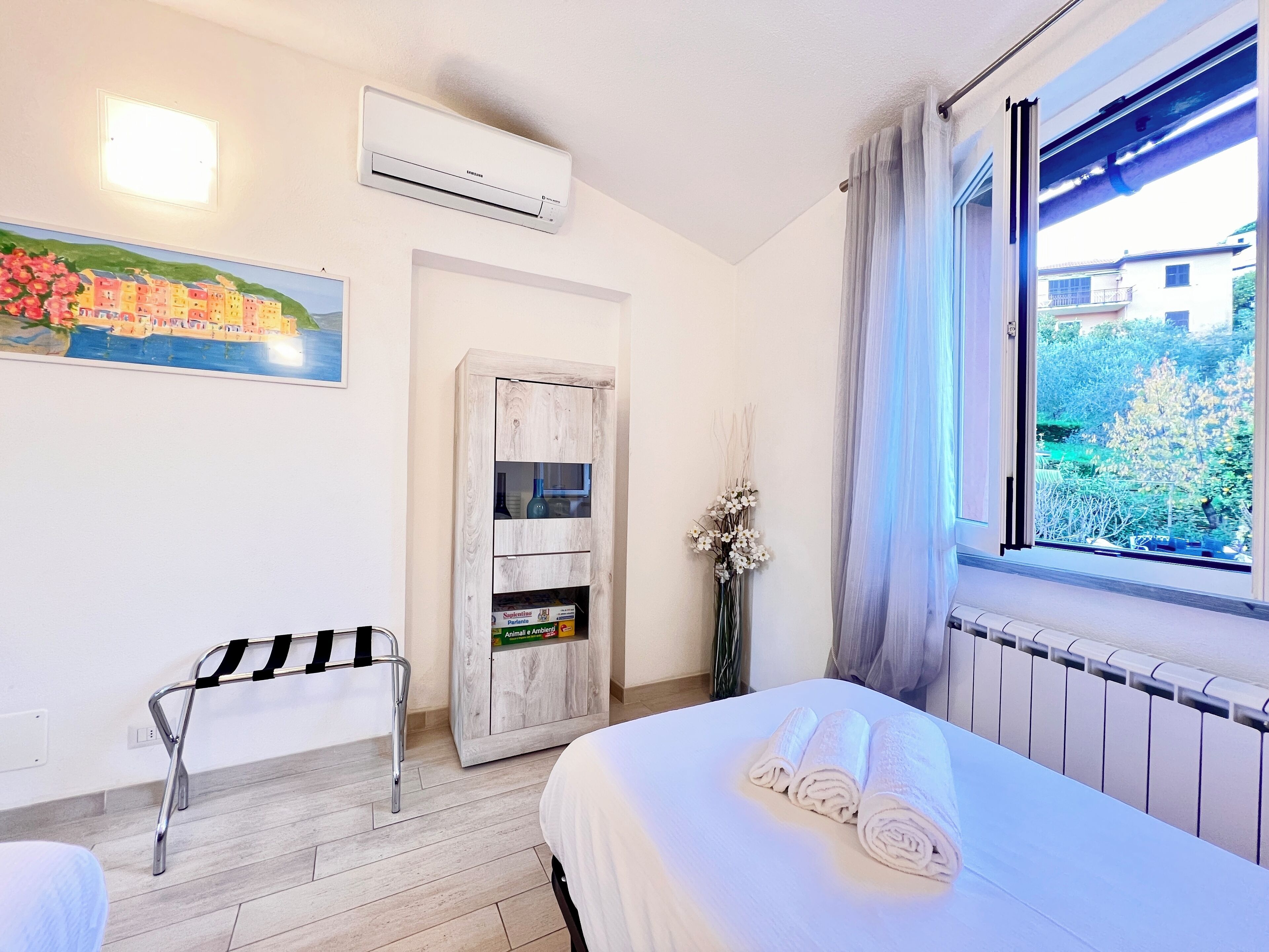 Hotelkamer met een bed, een stoel, een klein kastje, een airconditioning en een raam met uitzicht op groen.