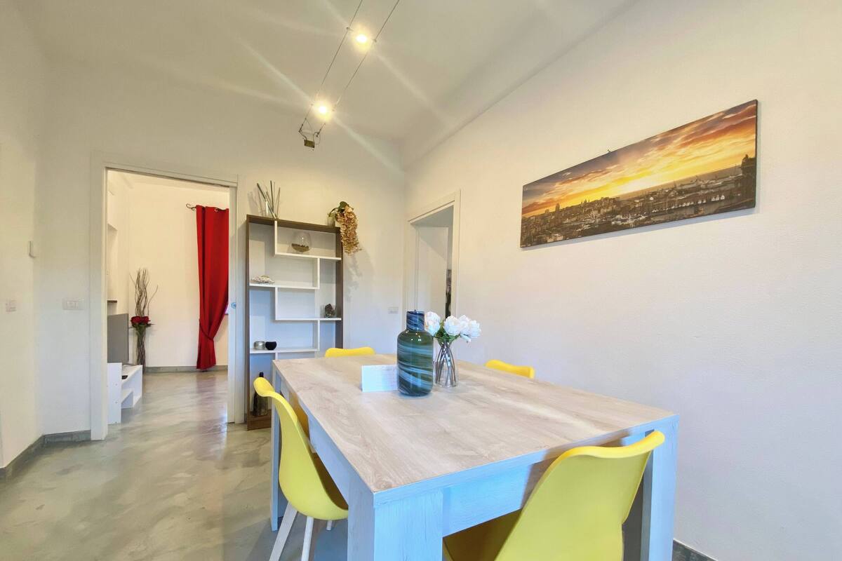 Un comedor con una mesa de madera y sillas amarillas, un jarrón grande con flores y un cuadro de un paisaje urbano en la pared.