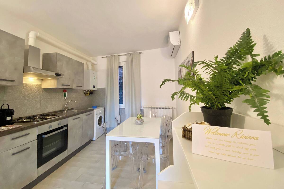 Una cocina moderna con estufa, horno y lavadora. Hay una mesa de comedor con sillas y una planta en maceta. Un letrero de bienvenida está en la pared.