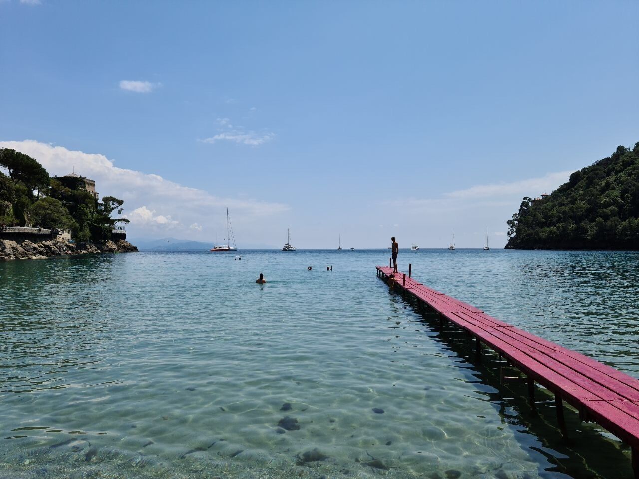 Un muelle rojo se extiende sobre aguas tranquilas, frente a una orilla despejada, rodeada de exuberante vegetación y veleros a lo lejos.