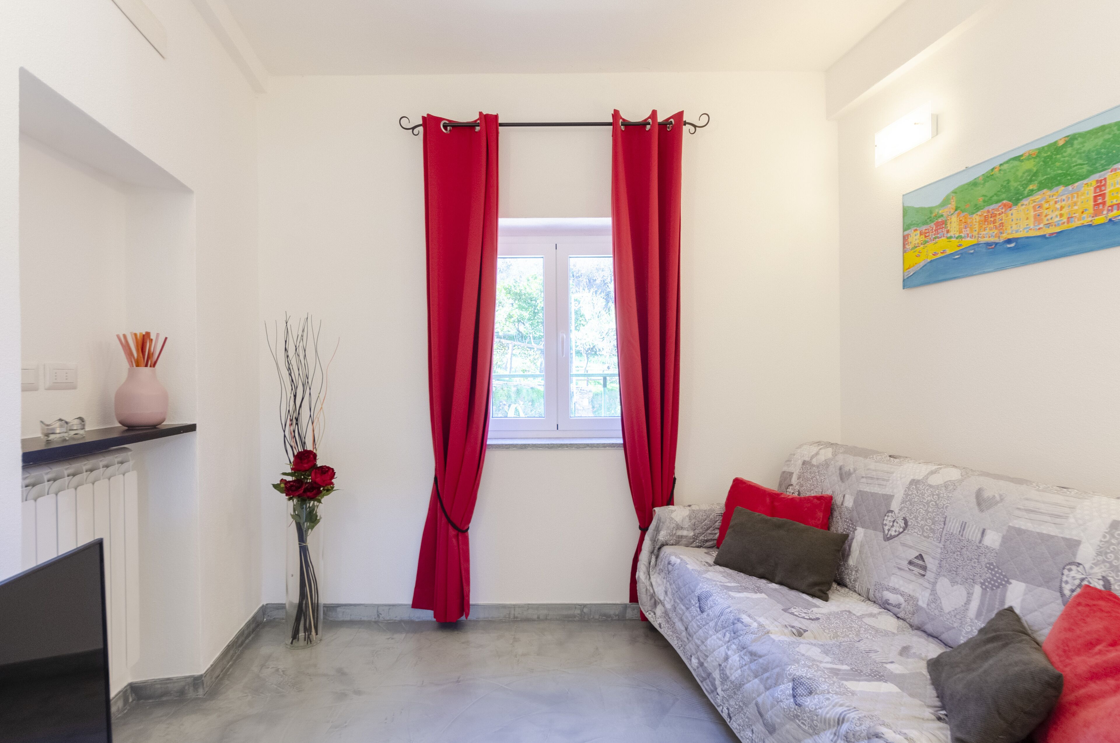 A room with a bed, a window with red curtains, a painting on the wall, and a vase with flowers.