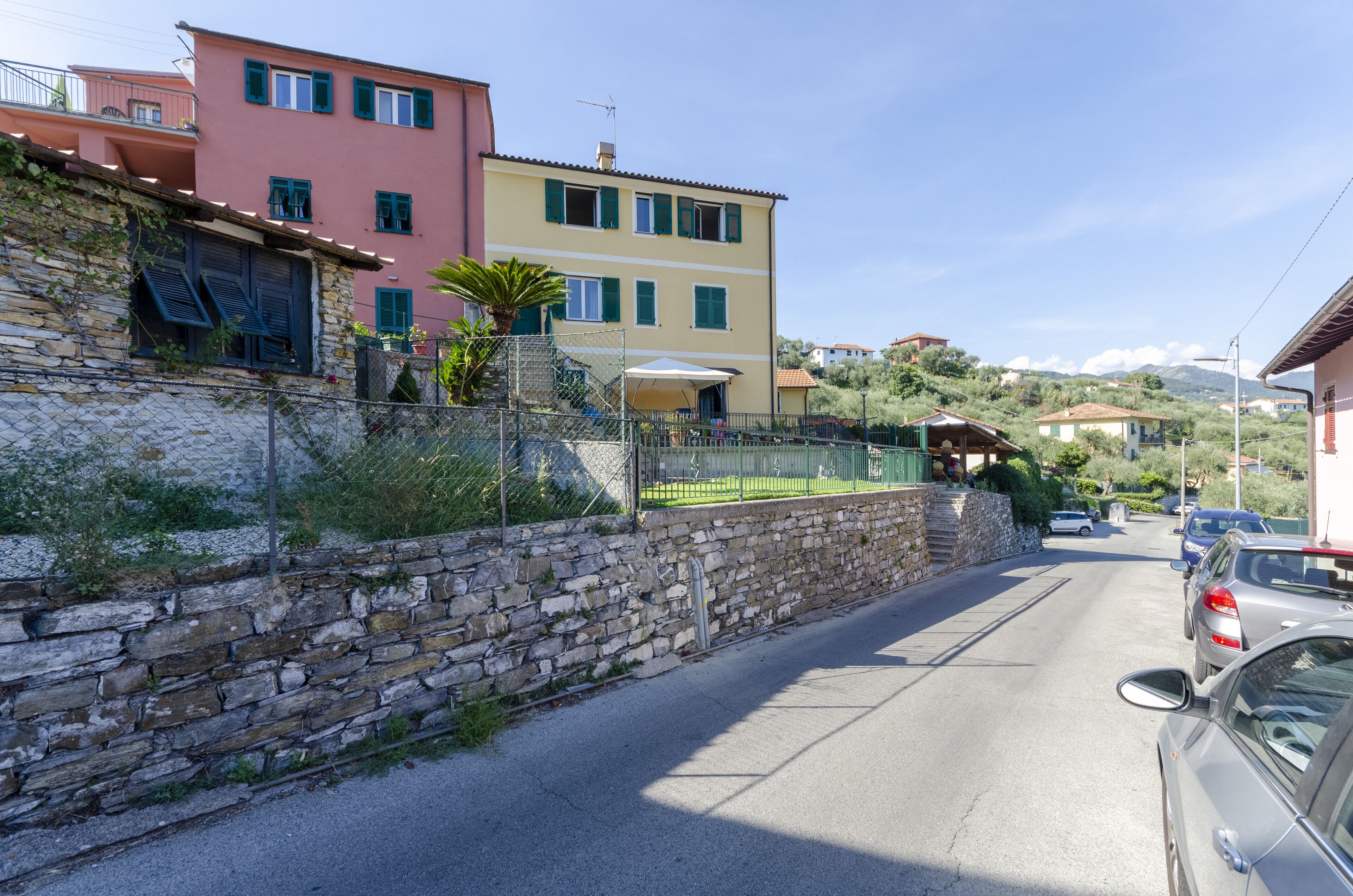 Uma rua estreita, com edifícios coloridos, uma parede de pedra e carros estacionados.