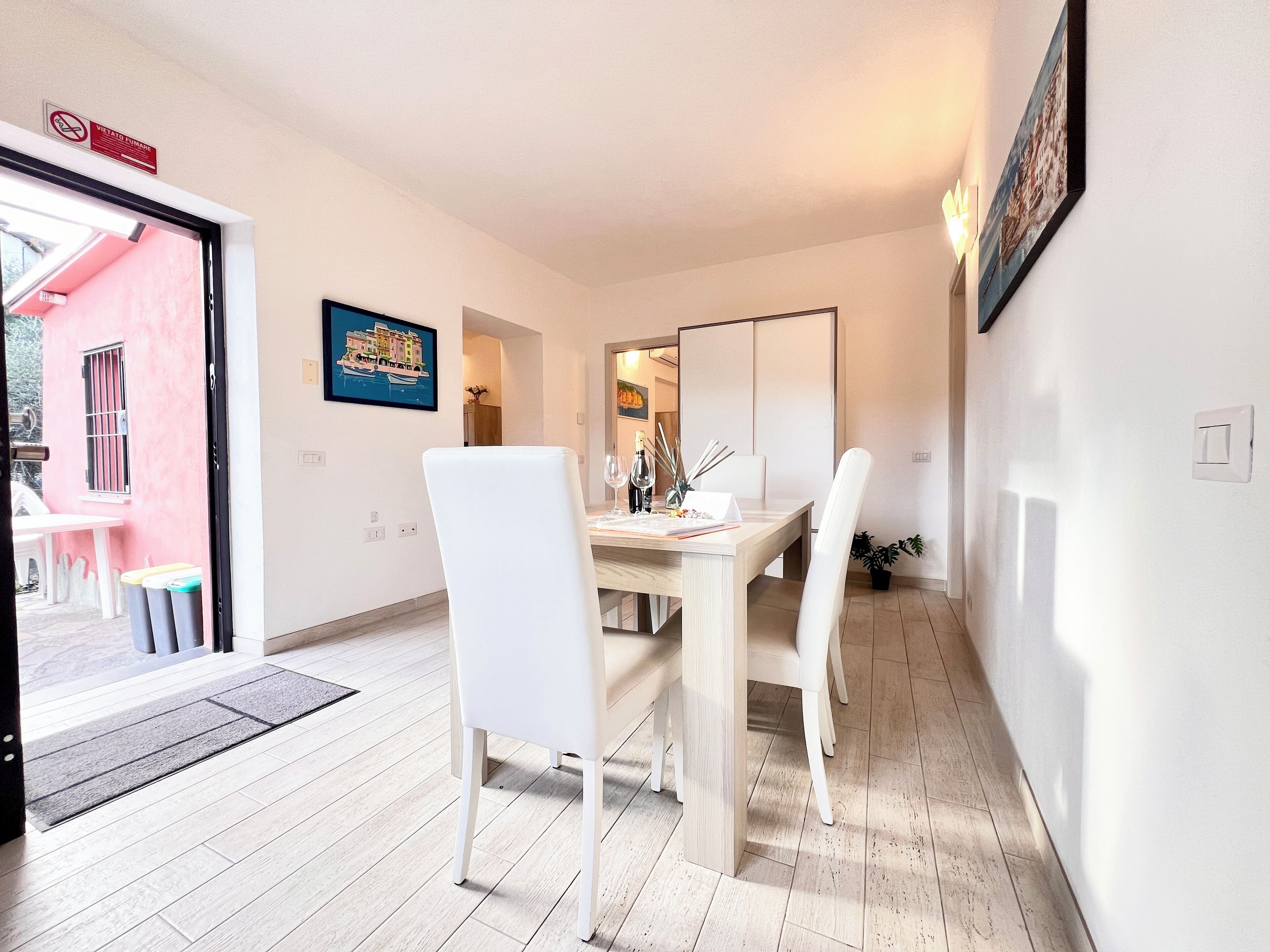 A dining area with a table set for four, white chairs, a painting on the wall, and a door leading outside.