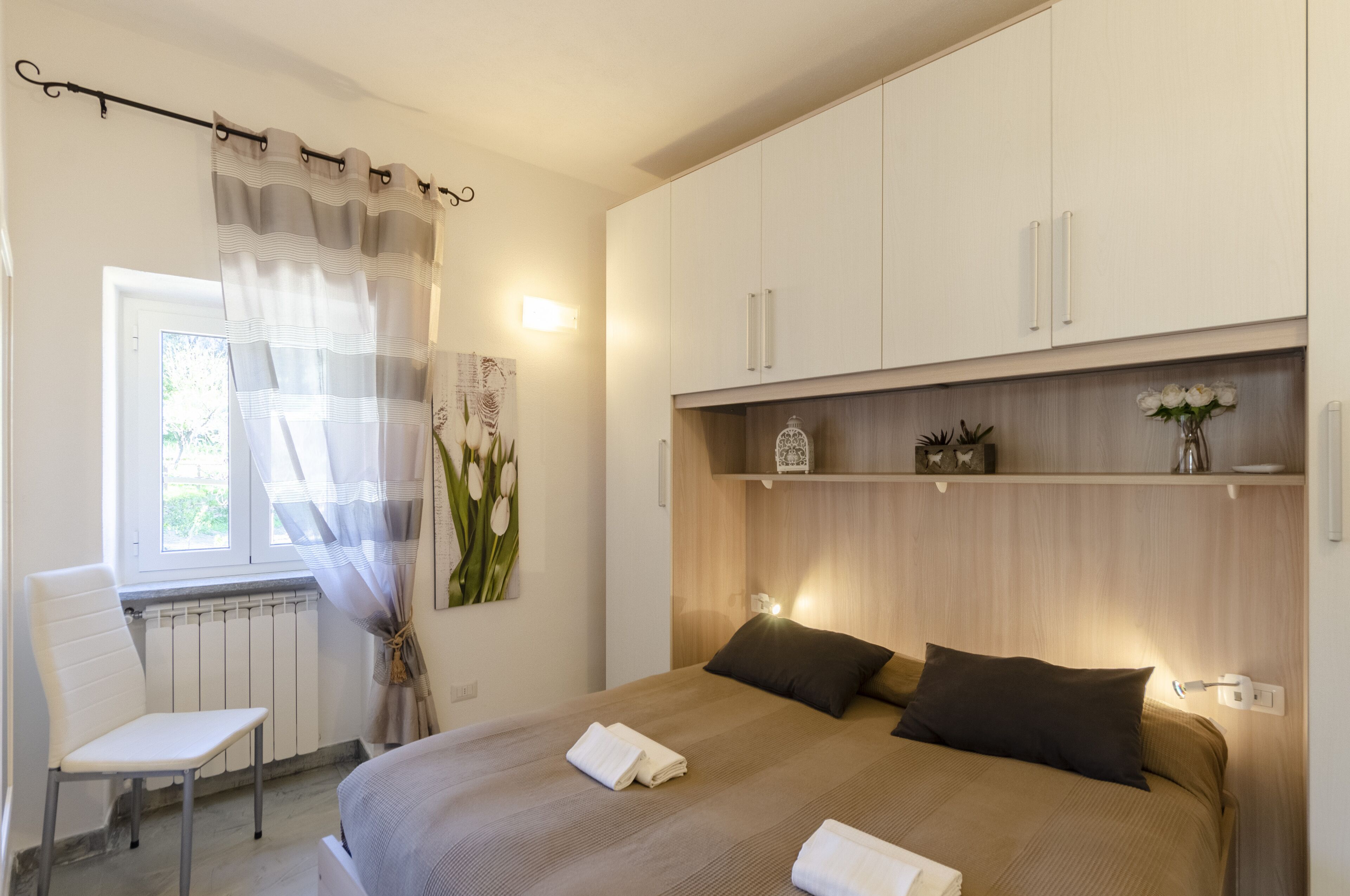 Un dormitorio con una cama, una silla, una ventana con cortinas, un radiador y una estantería con objetos decorativos.