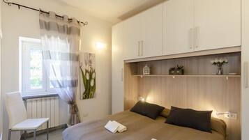 Un dormitorio con una cama, una silla, una ventana con cortinas, un radiador y una estantería con objetos decorativos.