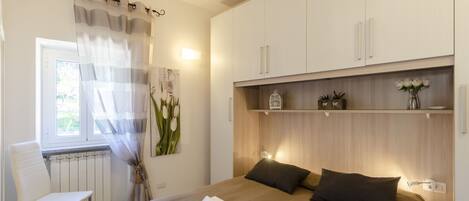 Un dormitorio con una cama, una silla, una ventana con cortinas, un radiador y una estantería con objetos decorativos.