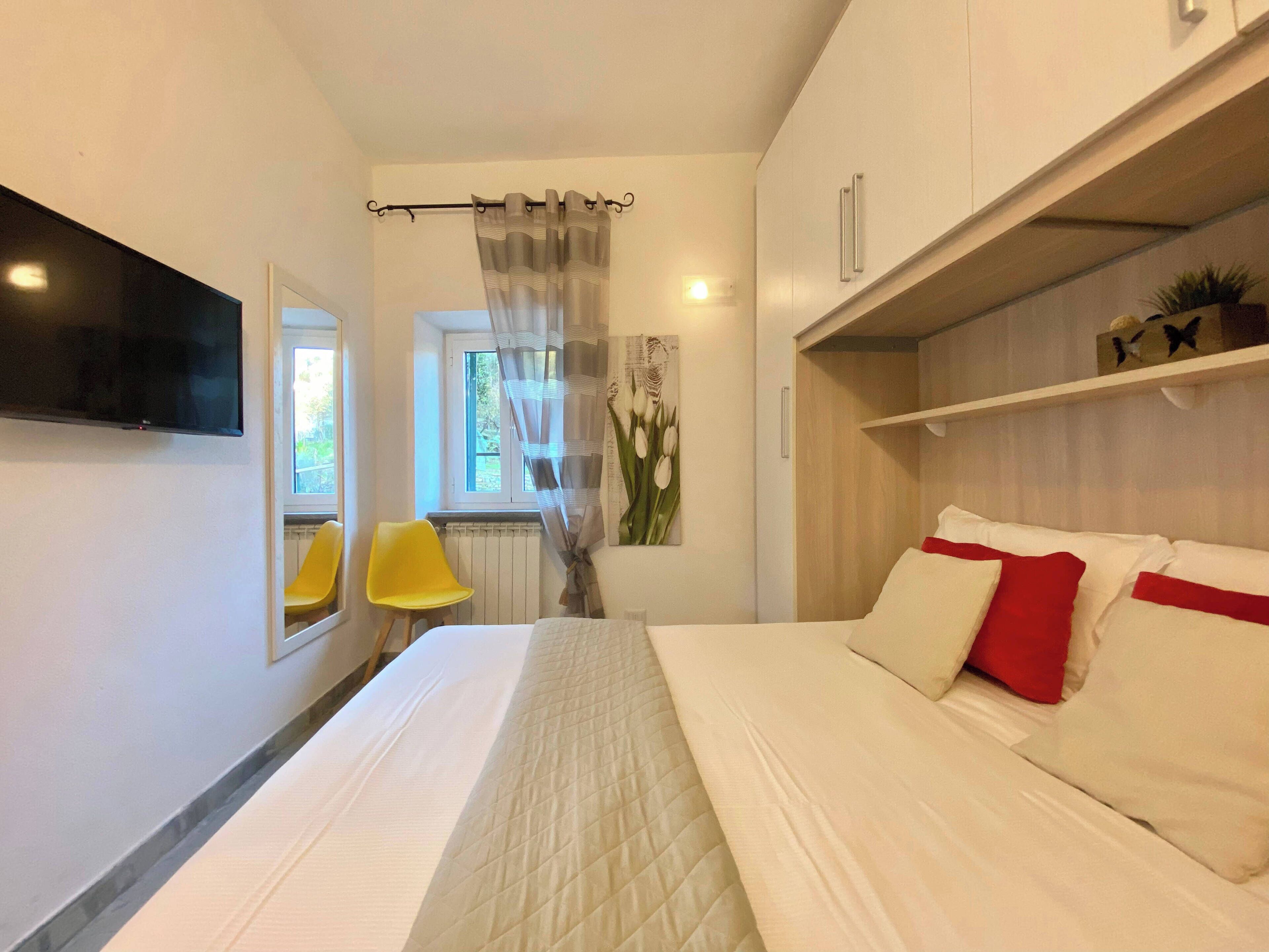 A bedroom with a bed, a TV mounted on the wall, a window with curtains, a chair, and a shelf with decorative items.