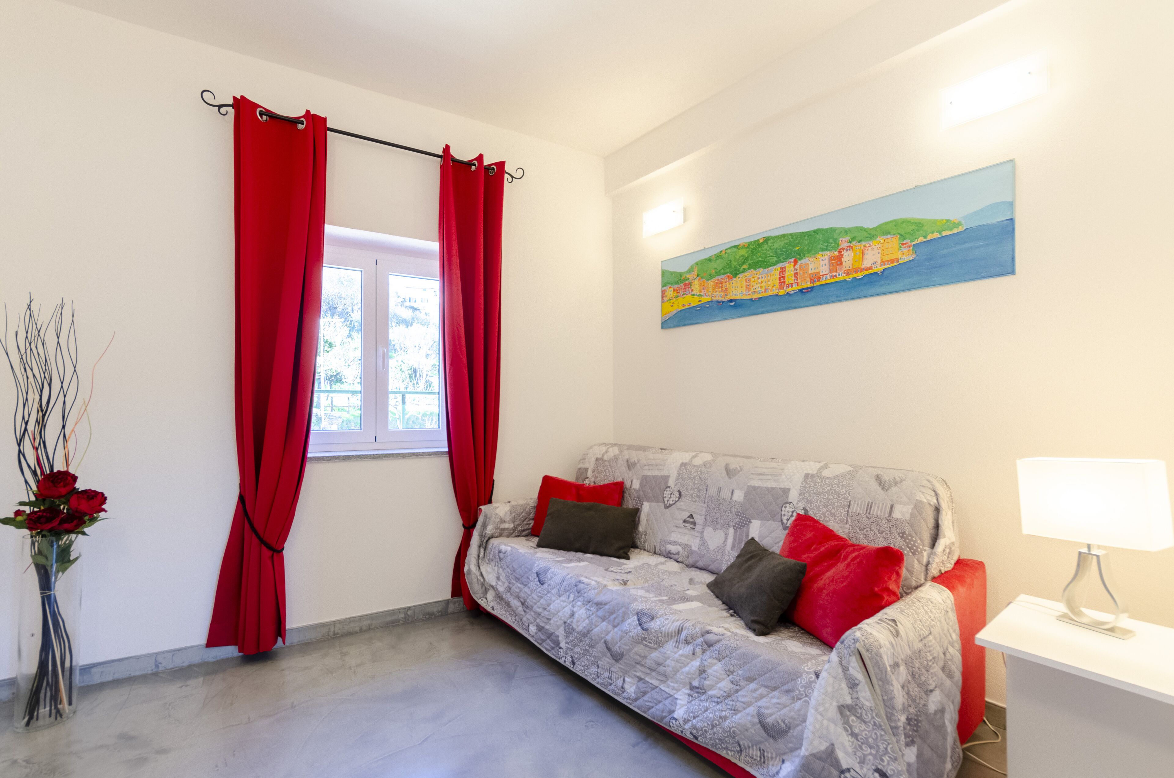 A room with a sofa, a window with red curtains, a vase with flowers, and a painting on the wall.