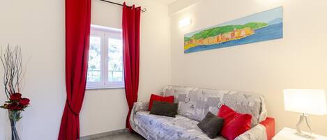 A room with a sofa, a window with red curtains, a vase with flowers, and a painting on the wall.