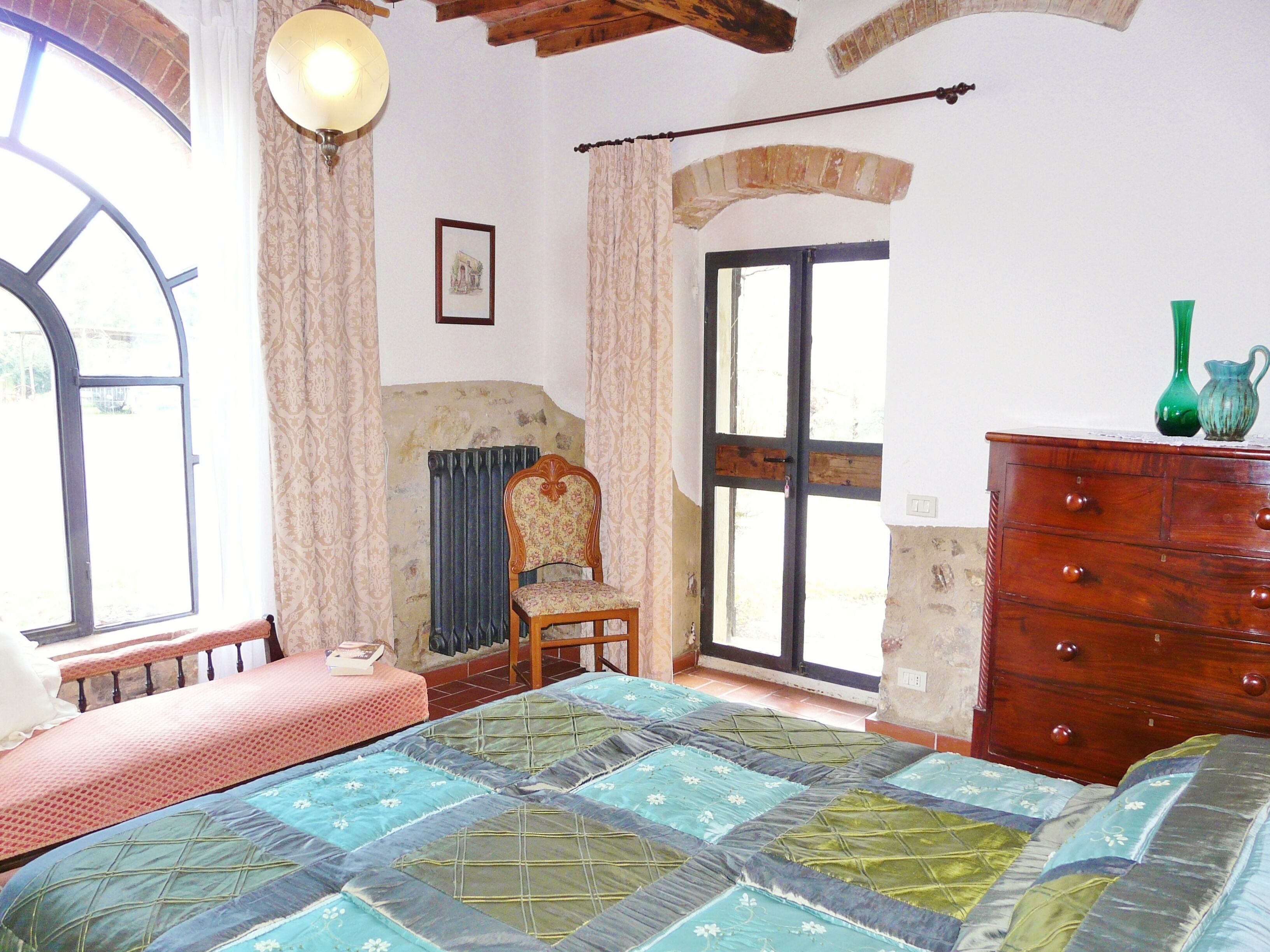Une chambre à coucher avec un lit, une commode, une chaise, un radiateur et une fenêtre avec des rideaux.