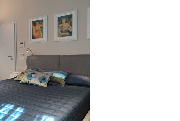 A bedroom with a bed, two framed paintings on the wall, and a bedside table with a lamp.